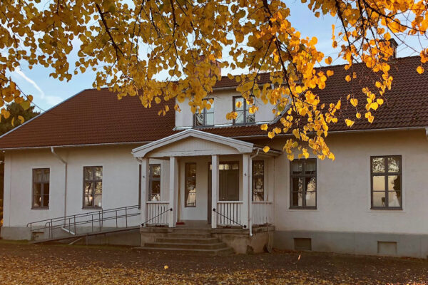 Vit bygdegårdsbyggnad med rött tegeltak står i höstsol, sedd framifrån. Gula löv hänger från ett träd och ramar in entrén med trappa, räcken och ramp; marken är täckt av fallna löv.