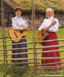 Två kvinnor i traditionella folkdräkter står bakom ett lågt trästaket och håller varsin akustisk gitarr, leende mot kameran. I bakgrunden syns en grön äng och en gammal träbyggnad. Text: “Foto: Strandroths fotografi”.