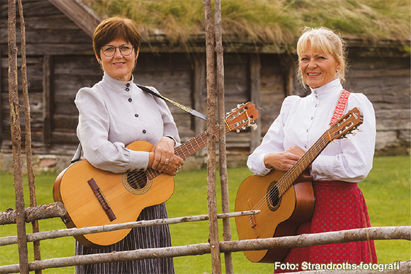 Två kvinnor i traditionella folkdräkter står utomhus och håller varsin akustisk gitarr, leende mot kameran. De står bakom en enkel träinhägnad med en äldre träbyggnad och gräs i bakgrunden. Text: ”Foto: Strandroths-fotografi”.