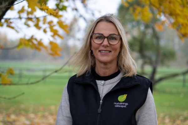 Kvinna med blondt hår och stora glasögon ler mot kameran utomhus. Hon bär svart väst med logga och texten “Hushållnings sällskapet”, framför suddig grön parkmiljö och gulnande höstlöv.