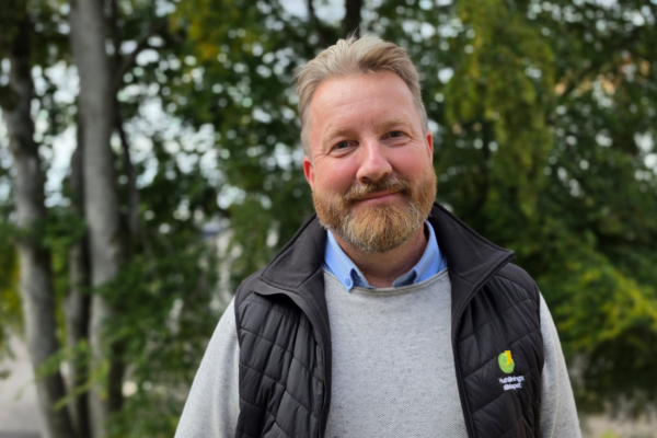 Skäggig man ler mot kameran och står utomhus framför suddiga träd. Han bär ljusgrå tröja, blå skjorta och svart vadderad väst med loggan och texten ”Hälsinglands Sparbank”.