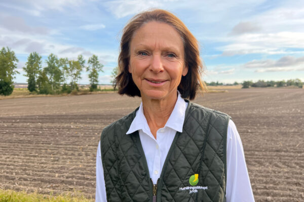 Kvinna i vit skjorta och grön, quiltad väst står och tittar mot kameran på en plöjd åker med träd och blå himmel i bakgrunden. Brodyr på västen: “Hushållningssällskapet Juridik”.
