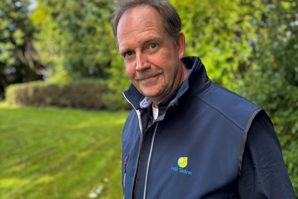 Man i mörkblå väst står utomhus och tittar mot kameran med ett svagt leende. Västen har logotypen och texten ”HIR Skåne”. I bakgrunden syns grön gräsmatta, träd och blå himmel.