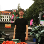 Man i svart t-shirt står och ler bakom ett marknadsbord med skålar av tomater och jordgubbar, gurka och zucchinis. I bakgrunden syns torgmiljö med träd, byggnader och rosa flaggor.