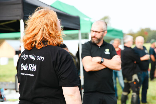 Kvinna med rött hår står med ryggen mot kameran och pratar med en man vid ett utomhusevent med tält. Text på tröjan: “Fråga mig om Goda Råd” och “till mjölk- och köttproducenter”.