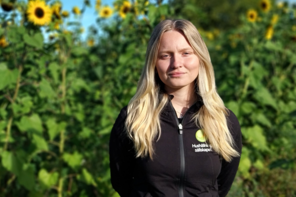 Kvinna med långt blont hår står och tittar mot kameran med lugnt uttryck, framför en grön solrosodling i soligt väder. Hon bär svart jacka med logotyp och texten ”Hushållningssällskapet”.