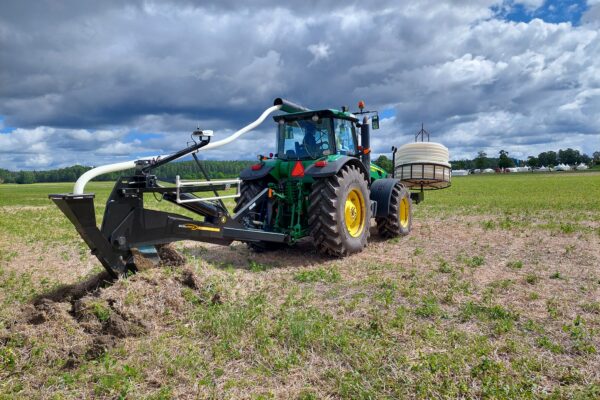Grön traktor med monterad täckdikningsplog gräver en smal fåra i ett fält, med vit slang som leds bakåt mot en stor slangrulle på släp. Mörka moln över landskap med skogsdunge i bakgrunden.