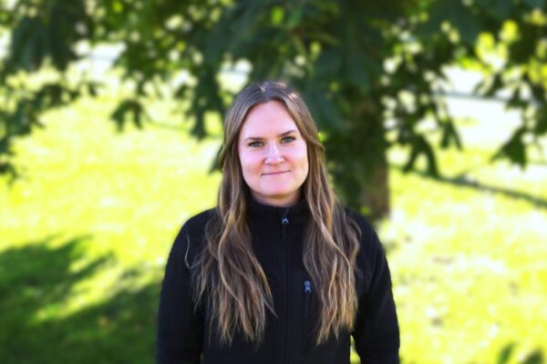 En kvinna med långt ljust hår står och tittar in i kameran med ett svagt leende, iklädd en svart jacka. Hon står utomhus i en solig grön park med träd och gräsmatta i bakgrunden.