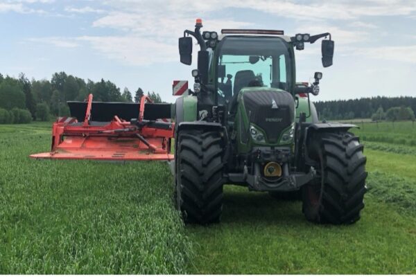 Grön traktor kör över en åker och slår gräs med ett rött slåtteraggregat framtill. Nyslaget gräs ligger i strängar på marken, med skogsridå och öppna fält i bakgrunden under molnig himmel.