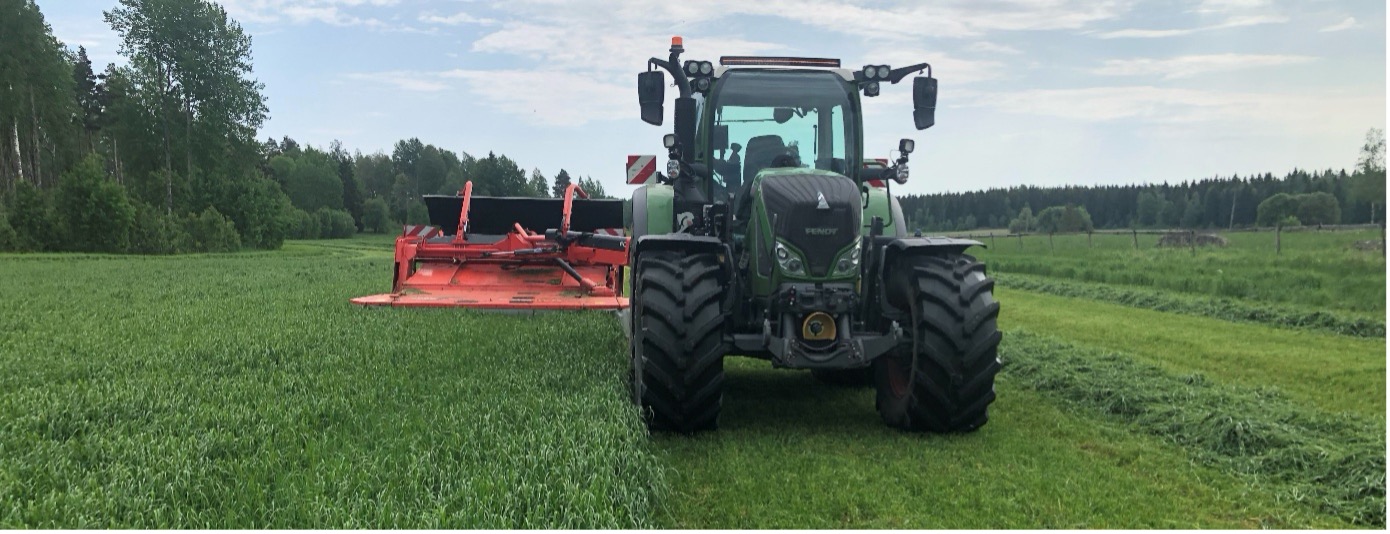 Grön traktor kör över en åker och slår gräs med ett rött slåtteraggregat framtill. Nyslaget gräs ligger i strängar på marken, med skogsridå och öppna fält i bakgrunden under molnig himmel.