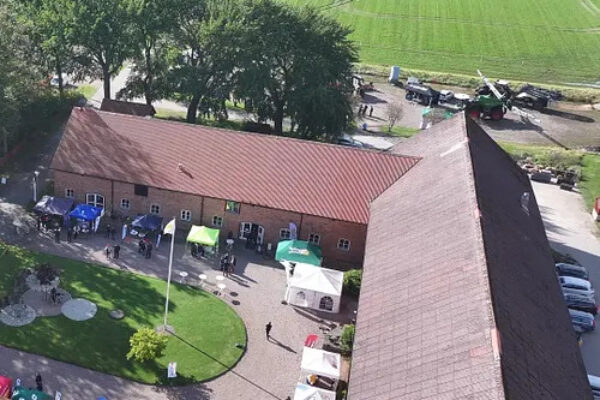 Flygvy över en gårdsplan vid en tegelbyggnad med rött tak, där besökare rör sig mellan vita tält och stånd. I bakgrunden syns gröna fält, träd och flera parkerade traktorer längs vägen.