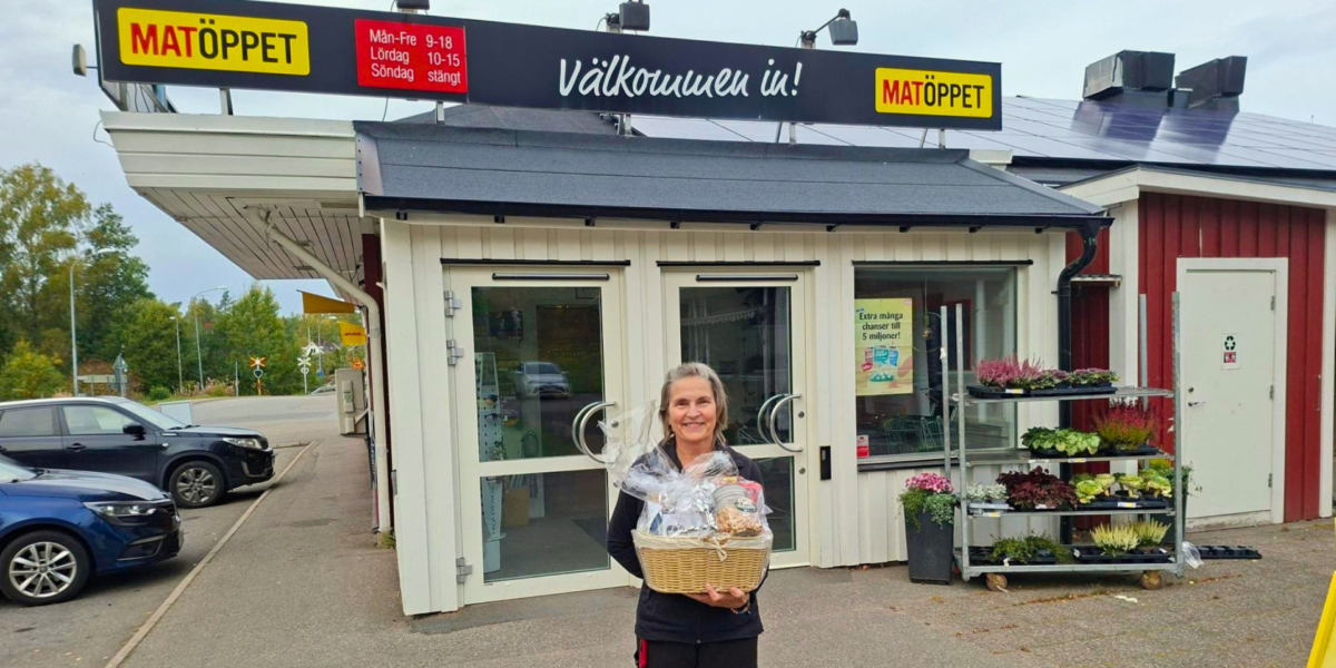 Kvinna står leende utanför butiken Matöppet och håller en inslagen presentkorg. Ovanför entrén syns skyltarna ”Välkommen in!” och öppettider: ”Mån–Fre 9–18, Lördag 10–15, Söndag stängt”; bredvid står blomvagnar.