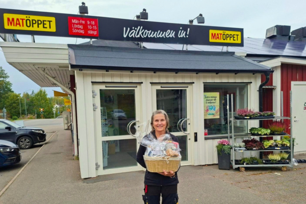 Kvinna står leende utanför butiken Matöppet och håller en inslagen presentkorg. Ovanför entrén syns skyltarna ”Välkommen in!” och öppettider: ”Mån–Fre 9–18, Lördag 10–15, Söndag stängt”; bredvid står blomvagnar.